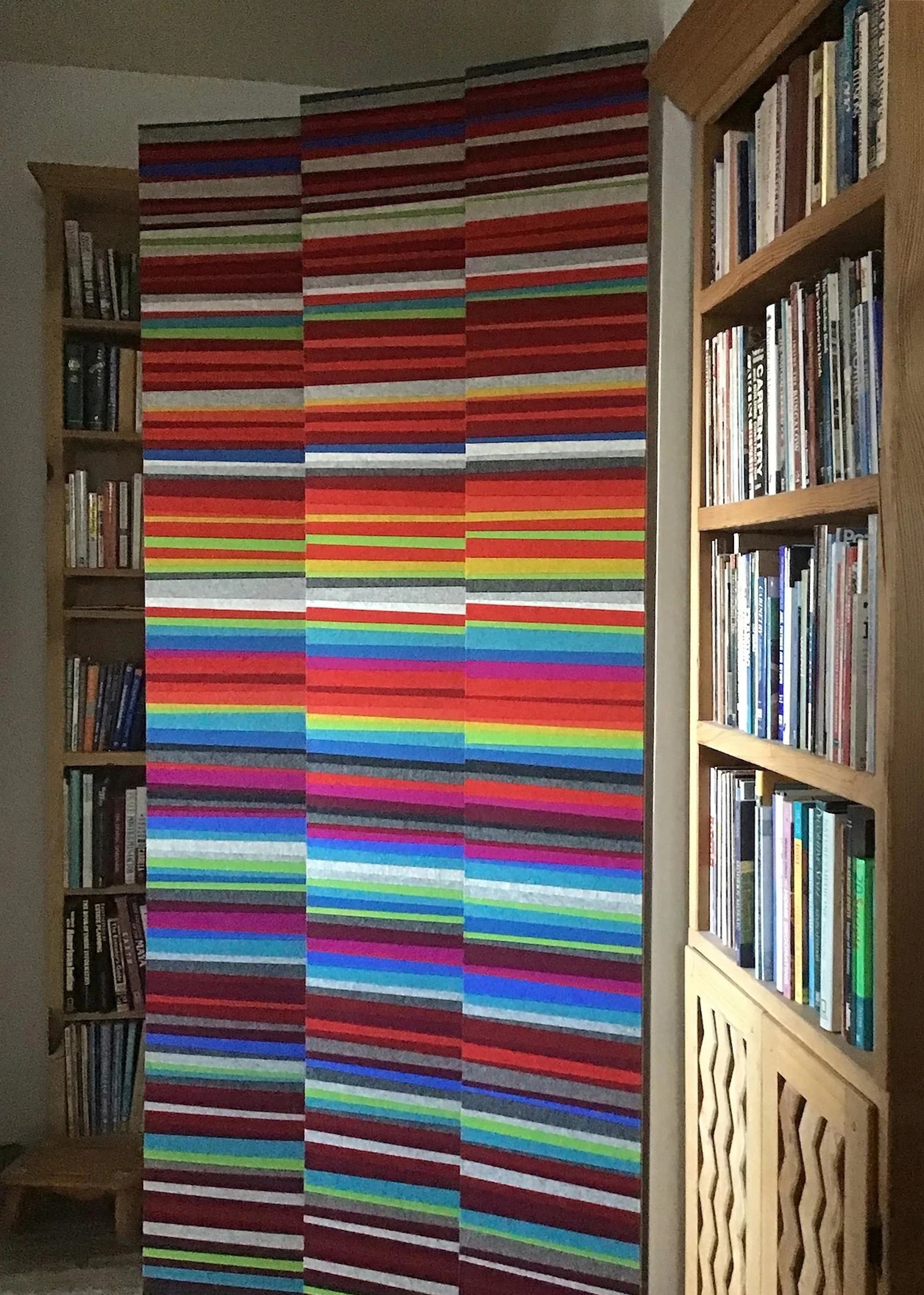 Three narrow panels stand next to some bookshelves. They are horizontal stripes of colorful felt.