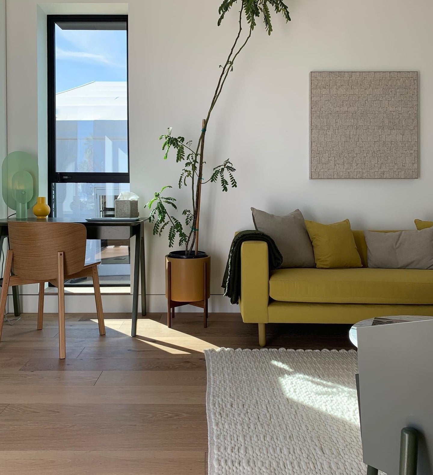 A sunny living room with modern furniture including a yellow couch. Hanging above the couch is a heathered gray felt wall panel with overlaying small block shapes.