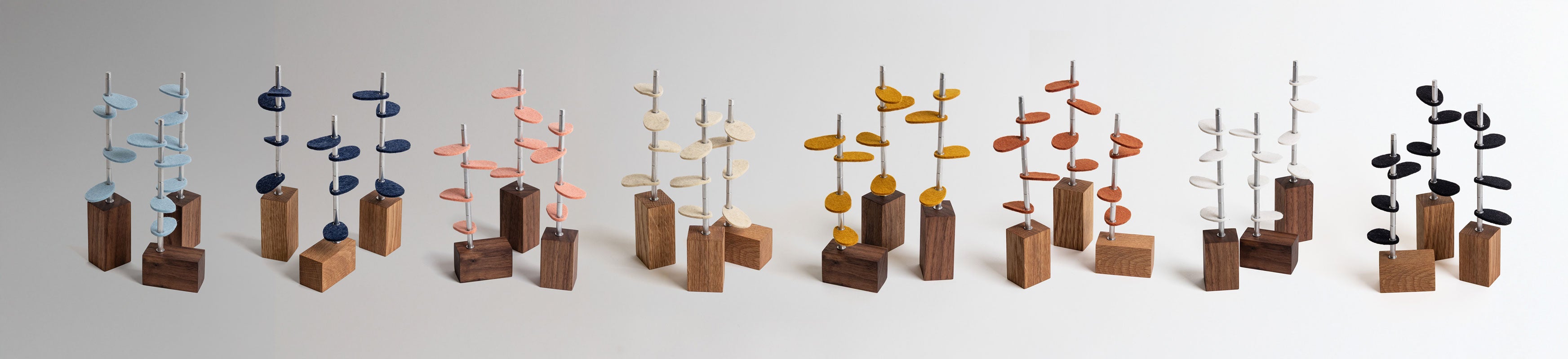 A long image of 8 sets of three sculptural objects of trees made of wood, metal stems, and colored wool felt leaves.