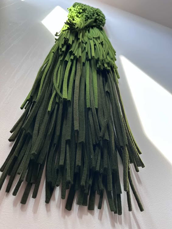 A handmade wall hanging or decorative sculpture made from many long, narrow green strips bundled together at the top, photographed against a plain white wall with bright sunlight and shadows.