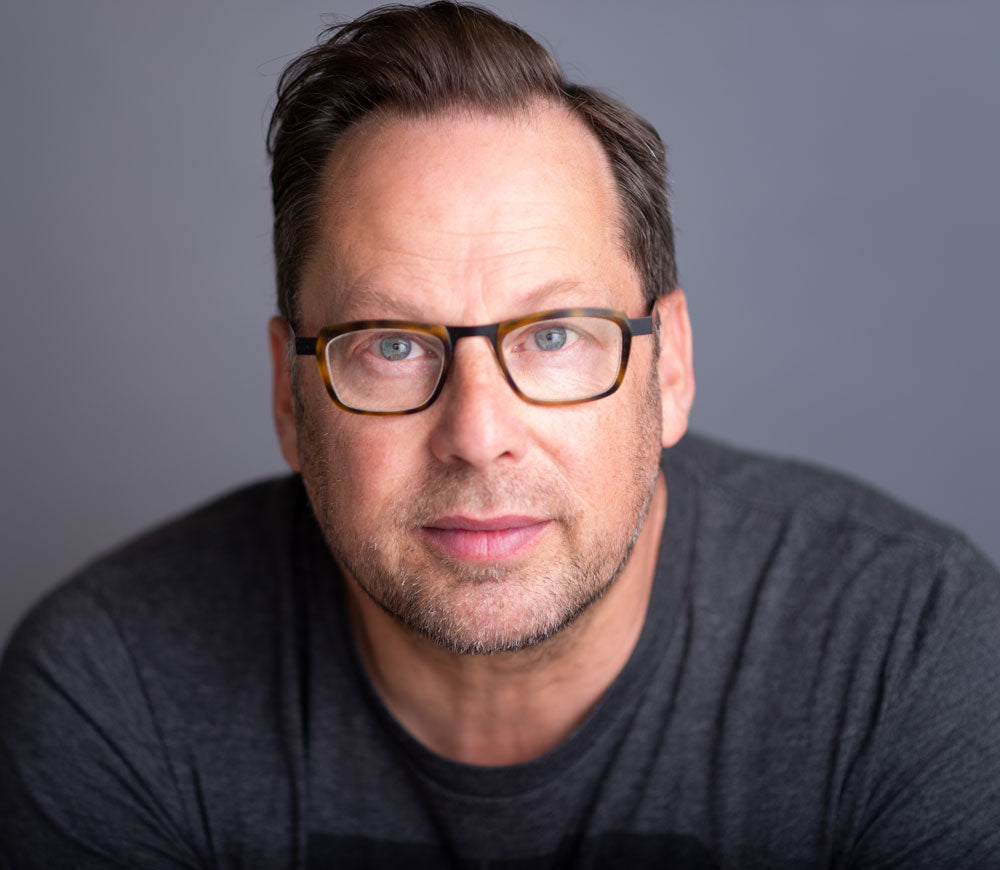 A white man with brown hair, tortoiseshell glasses and a short beard looks right at the camera.