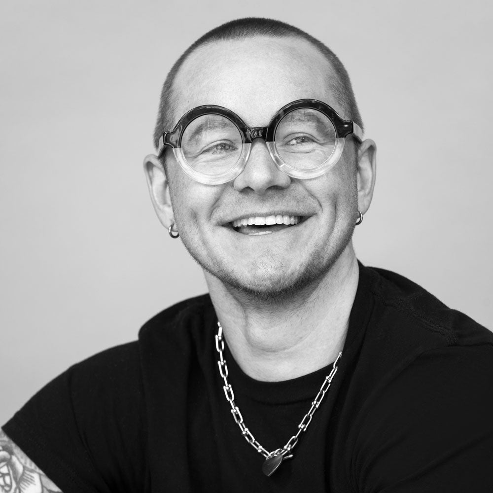 Black and white portrait of a person wearing glasses and a necklace on a plain background