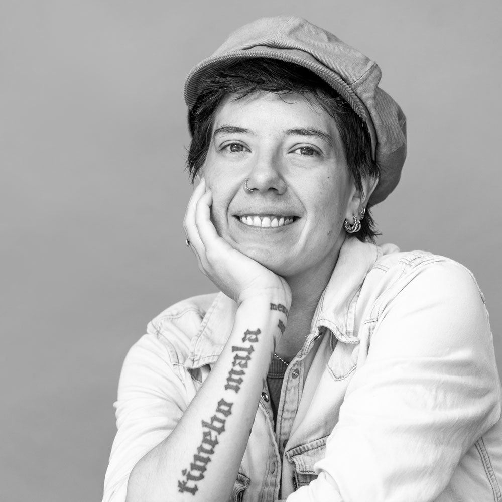 Black and white portrait of a person wearing a cap and denim jacket with visible tattoos.