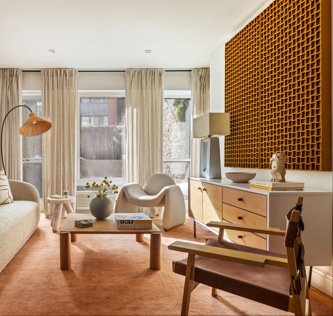 A modern living room interior with a beige textured sofa, matching armchair, wooden coffee table, terracotta rug, and a large mustard colored felt geometric wall panel on the right.