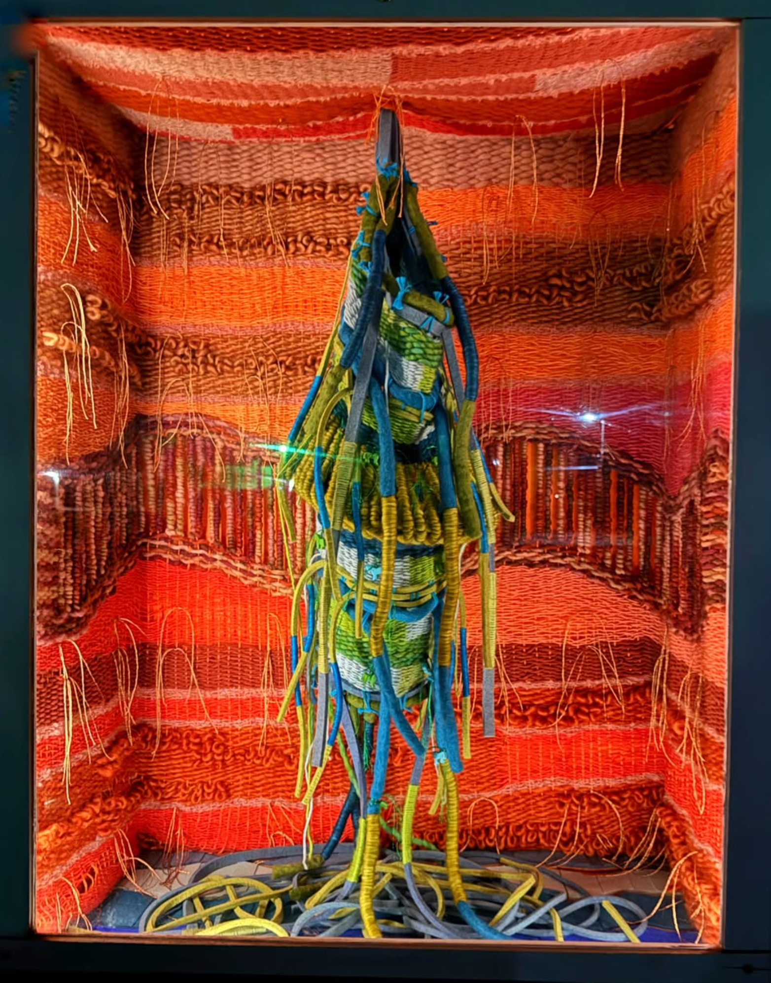 A colorful textile art piece or sculpture displayed inside a box-like structure, featuring dense woven and knotted patterns in orange, red, blue, yellow, and green, with long hanging strands and a central vertical form.