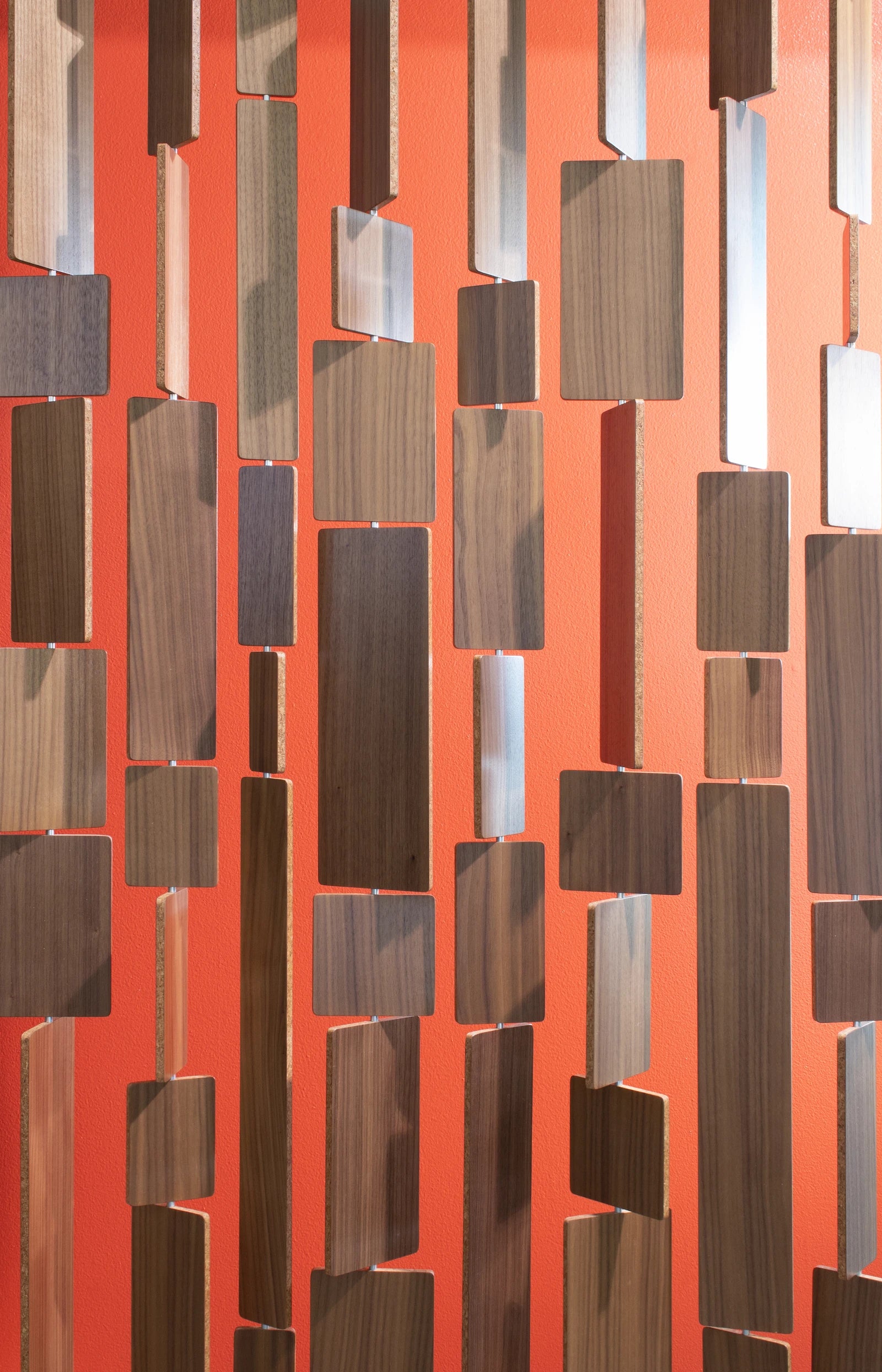 A close up of Century Screen room divider which has tiles of walnut veneer with a cork center hung in various lengths. Hanging in front of a bright orange wall.