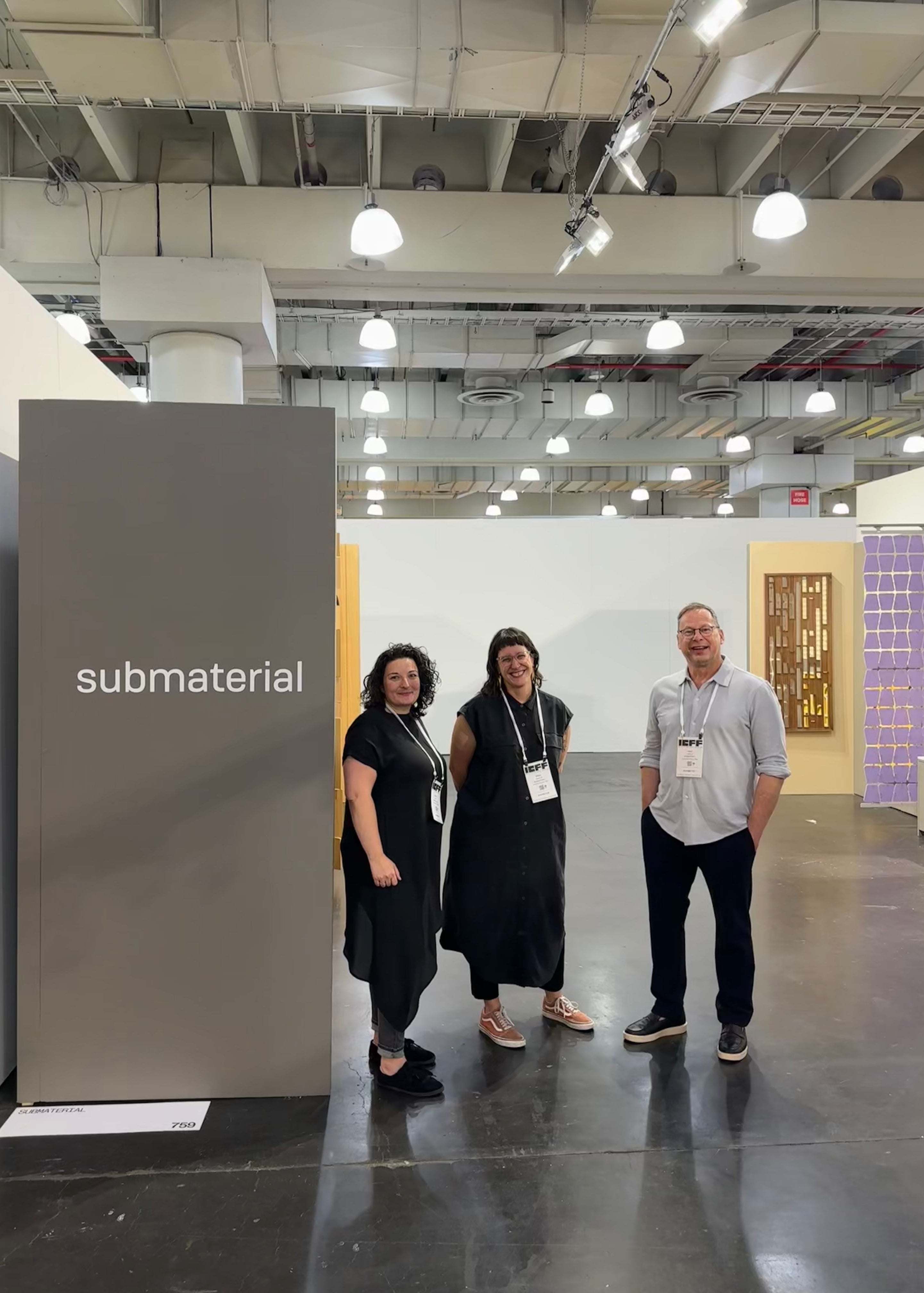 Two women with dark hair dressed in black and a man with a gray shirt and black pants stand in a tradeshow booth. he gray wall to the left says the word Submaterial.
