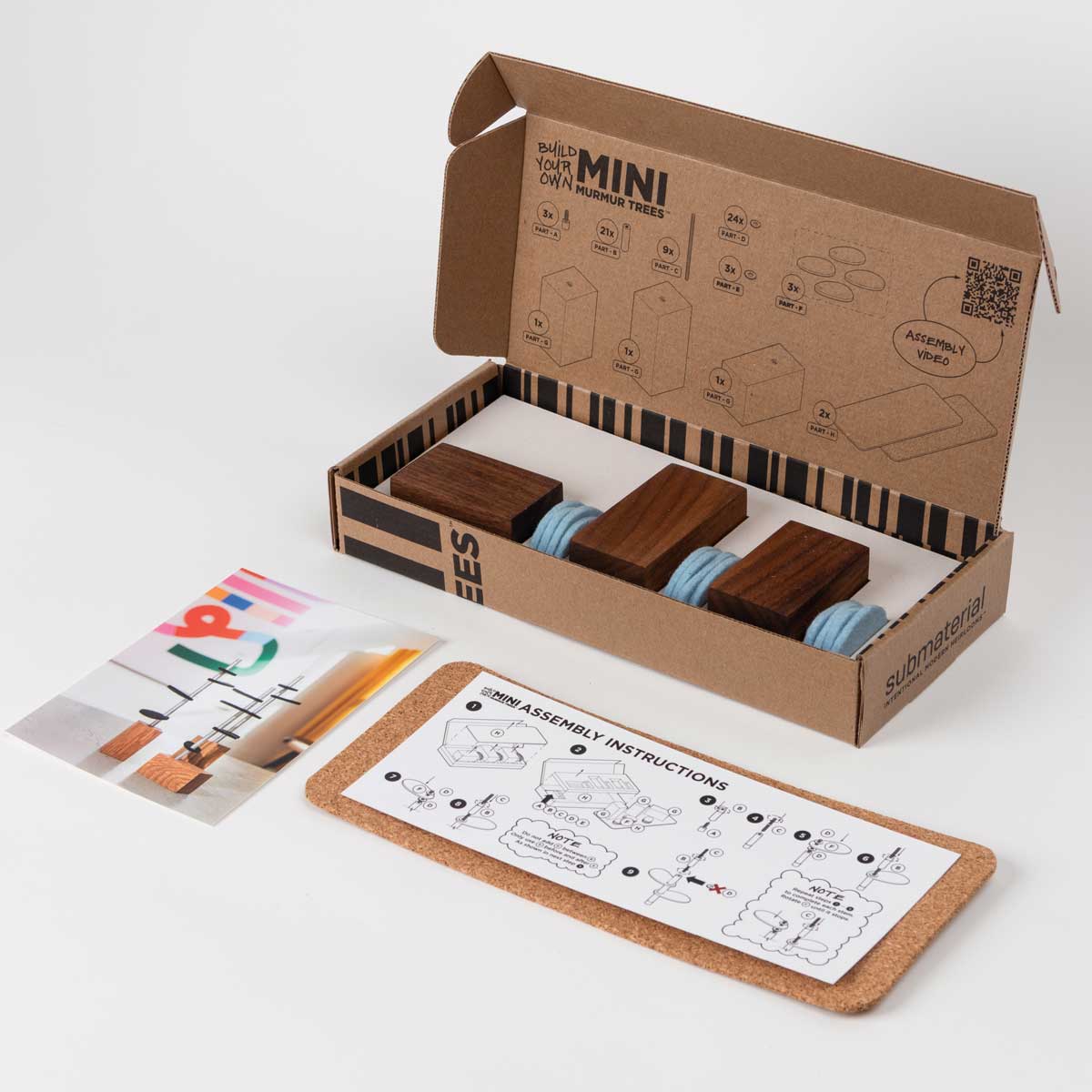 An open cardboard and black flat box with the wood bases and felt leaves of a murmur tree set. Laying infront of the box is a cork surface pad, a photo card and an instruction card.