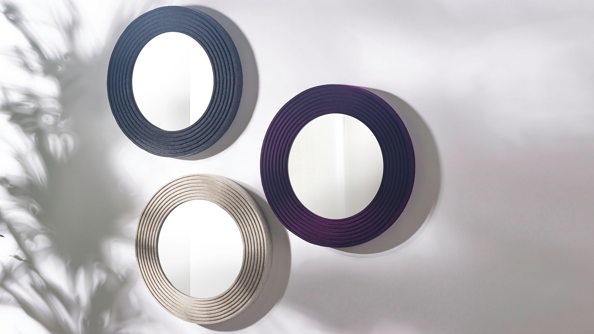 Three small round felt framed mirrors hang on a white wall. They are a dark heathered gray, an eggplant, and a light heathered tan.