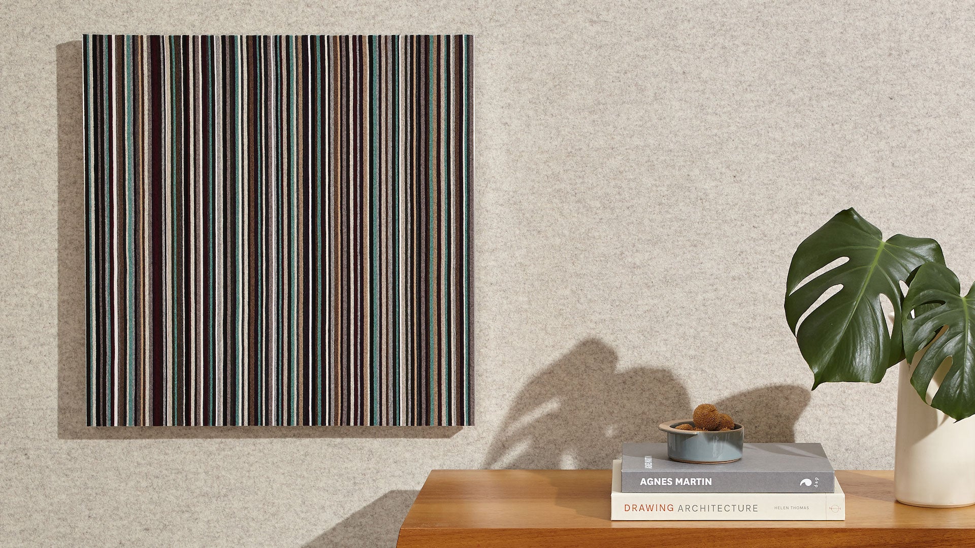 Modern felt wall art with vertical stripes on a gray felt wall next to a wooden table with books and a plant.
