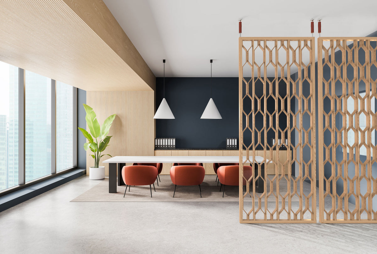A modern interior space with a tall light wood geometric screen partition on the right, a long white table, four orange-red upholstered chairs, white pendant lights, a dark navy accent wall, and large windows on the left.