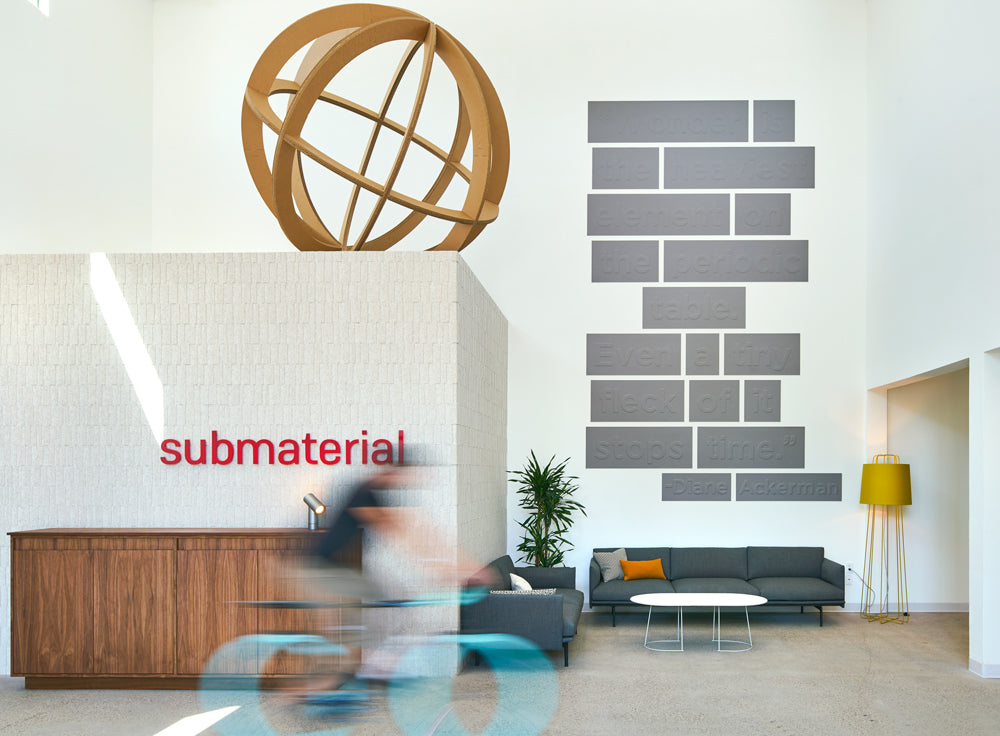 A lobby space with a textured white wall and wood cabinet at left. On top of the wall, a giant cardboard orb sits. At the right is a lounge area with modern gray couches and a white coffee table. The wall above the couches has a quite in gray. A blurred figure of someone riding a bike.