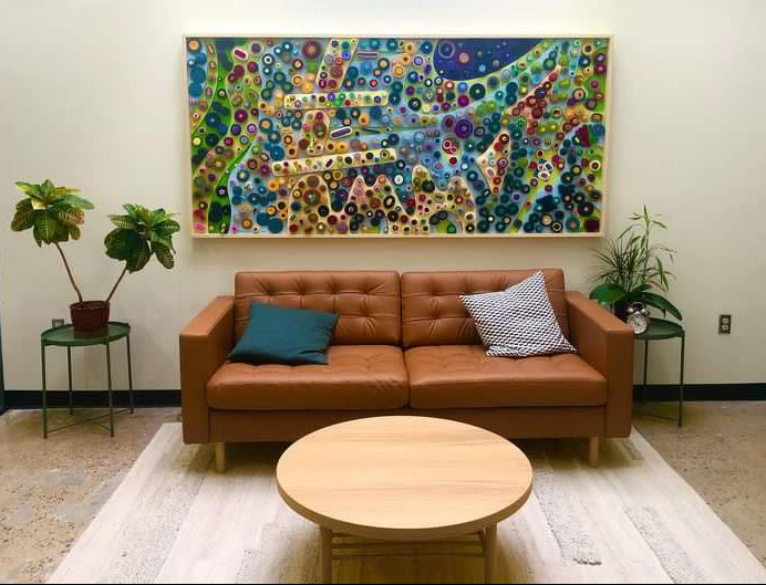 A large, vibrant, multicolored circular abstract wall art piece hangs on a white wall above a brown leather sofa in a modern living room setting with a round wooden coffee table and potted plants.