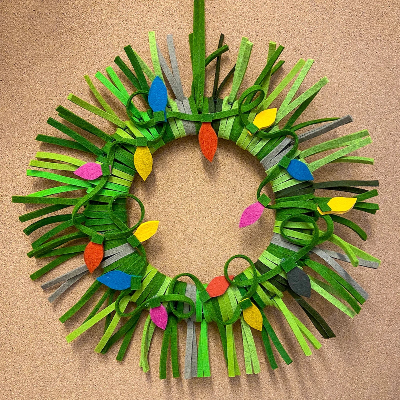A holiday wreath made of thin strips pof green and gray wool felt. A strand of lights also made of felt is attached to the front.