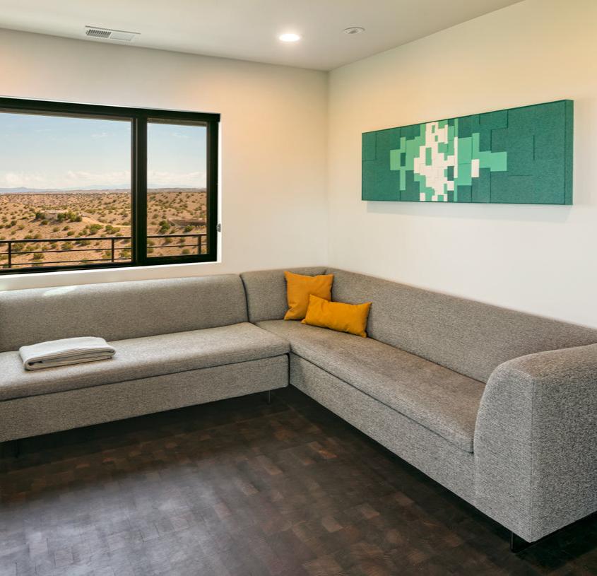 A bright living room with a sleek L-shaped couch. At right a rectangular felt wall panel in blocks of green and whites. To the left a large window showing the scrubby high desert. 