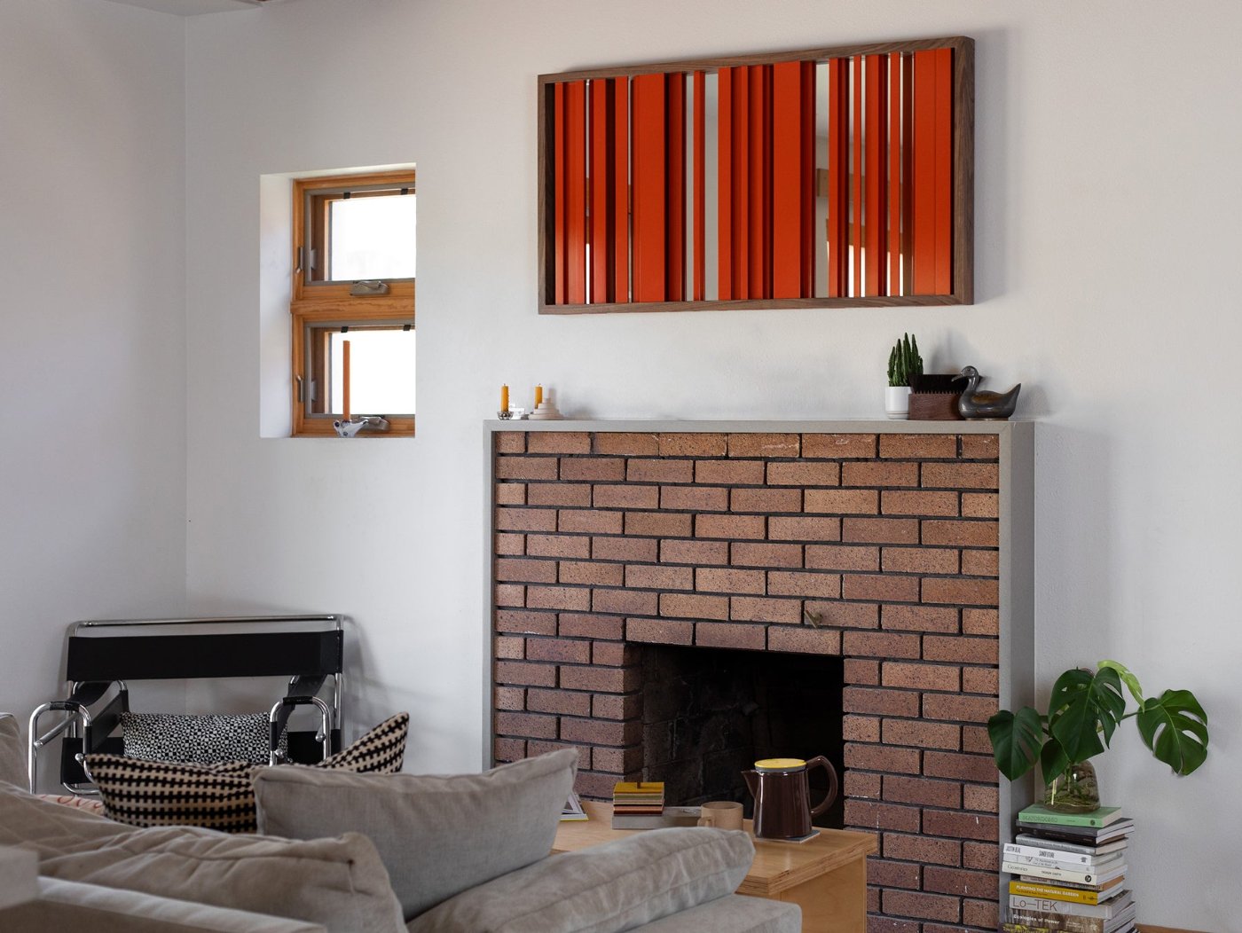 A cozy living area with a couch, chairs, and a brick fireplace. Above the mantle hangs a decorative mirror. In a wood frame, the mirrored glass in attached to the back while bars of range metal obscure the mirror.