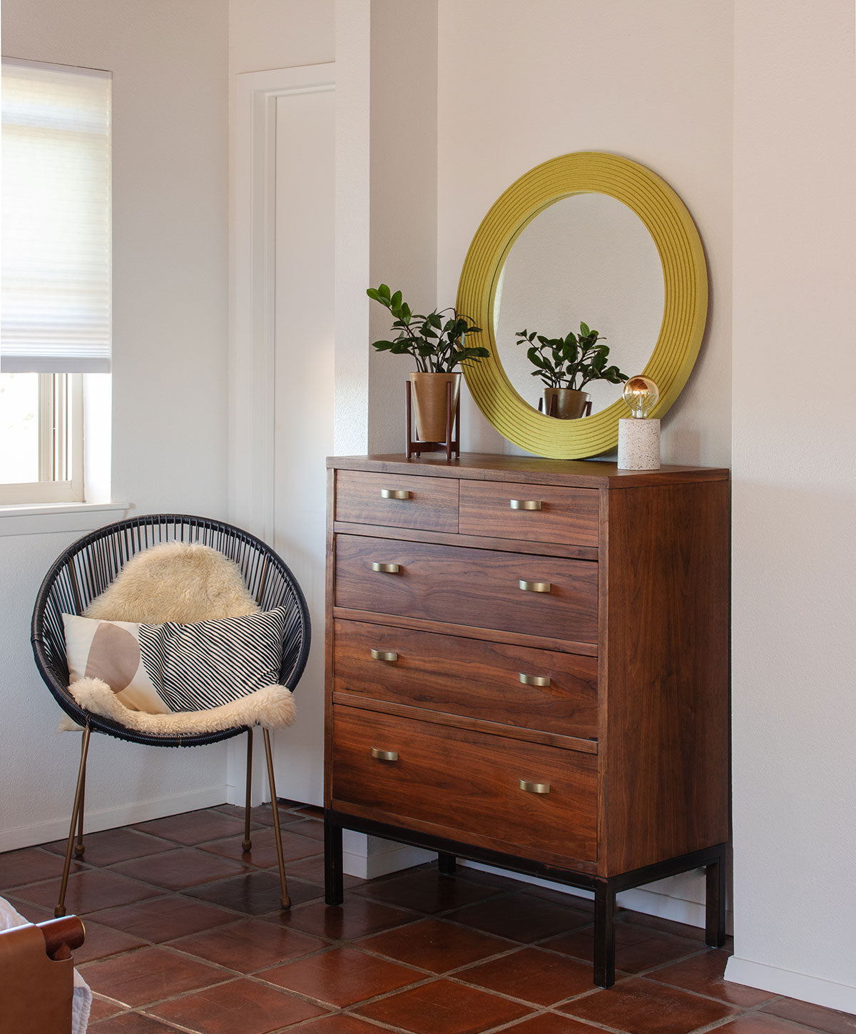 The corner of a bedroom with a thin black chair and a dresser. On the dresser sits a chartreuse round mirror made of alternating height strips of wool felt. 
#color_chartruese