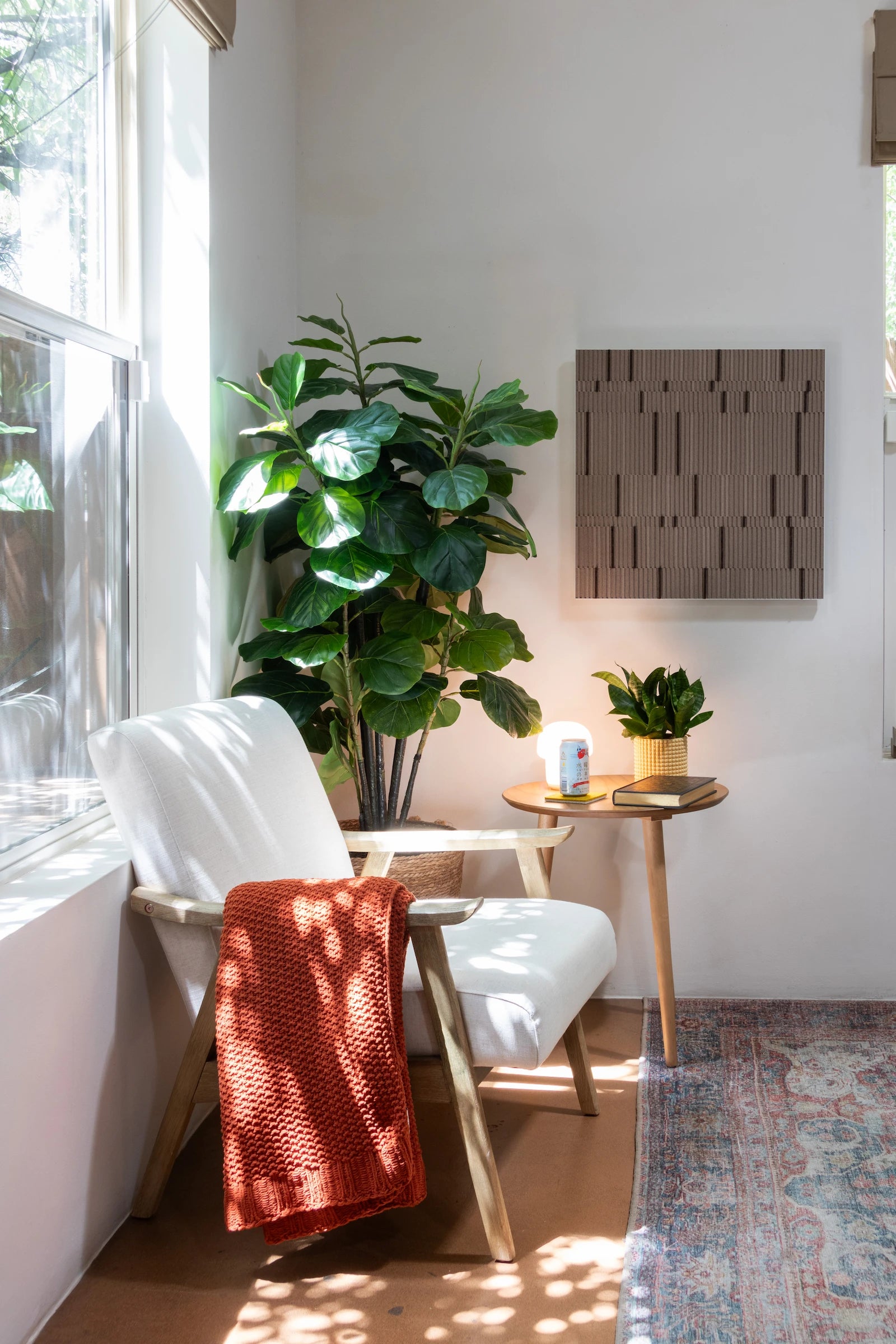Circadian No. 1 wall art piece of tan rows of alternating height felt with random bands of smoked cork in a square white metal cradle. It is hung on a white wall in a living room space with a large plant, side table and cozy chair. 