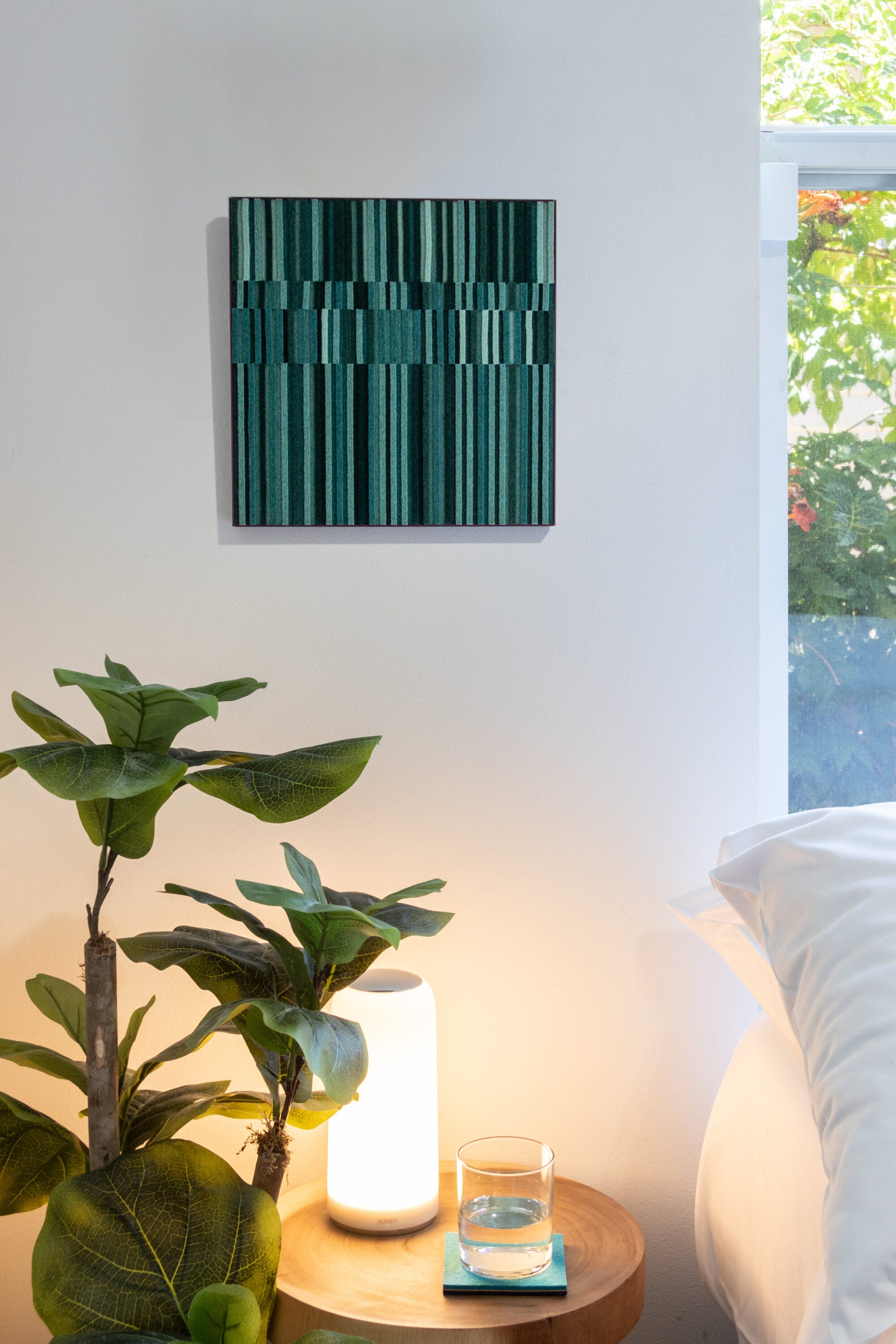 A small wall art piece of rows of alternating height felt in various tones of green in a violet colored metal cradle. Here Circadian No. 2 is hung on a white wall next with a bed with a side table and plant.