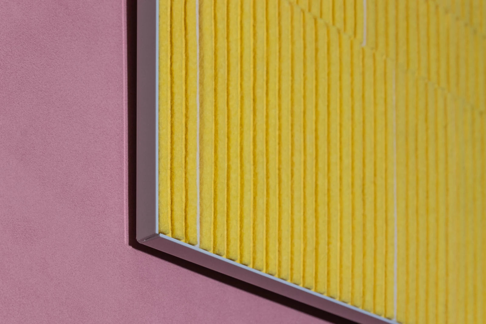 A closeup of a corner of a felt wall art piece in a white metal cradle. The piece is alternating heights of bright yellow felt with a couple of white strips seen at the lower height. The piece is seen against a pink wall.