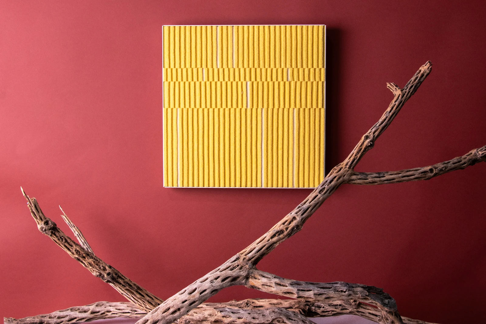 A felt wall art piece in a white metal cradle hangs on a red wall. The piece is alternating heights of bright yellow felt with a couple of white strips seen at the lower height. Under the piece are dried cholla branches.