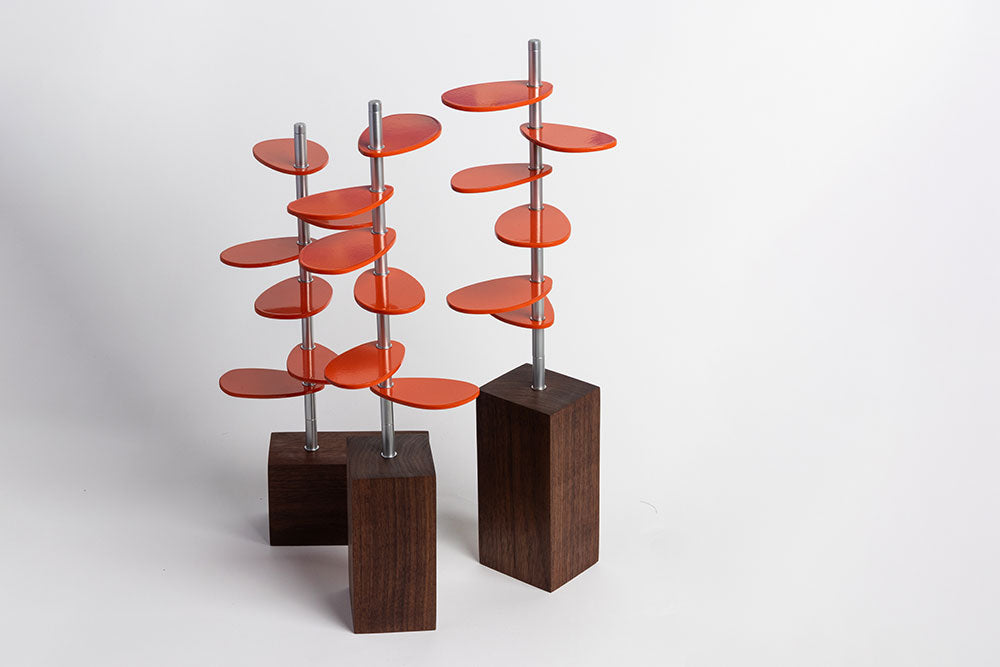 A straight on shot of Murmur Trees. Three sculptural objects with walnut wood block bases, brushed metal stems and orange powder-coated oval leaf shapes. #color_orange metal leaves