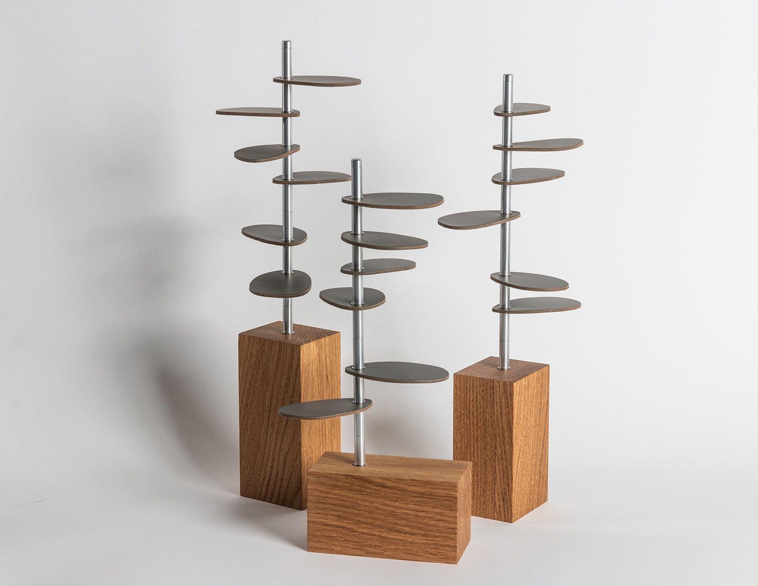 A straight on shot of Murmur Trees. Three sculptural objects with white oak wood block bases, brushed metal stems and gray leather oval leaf shapes. #color_gray leather leaves