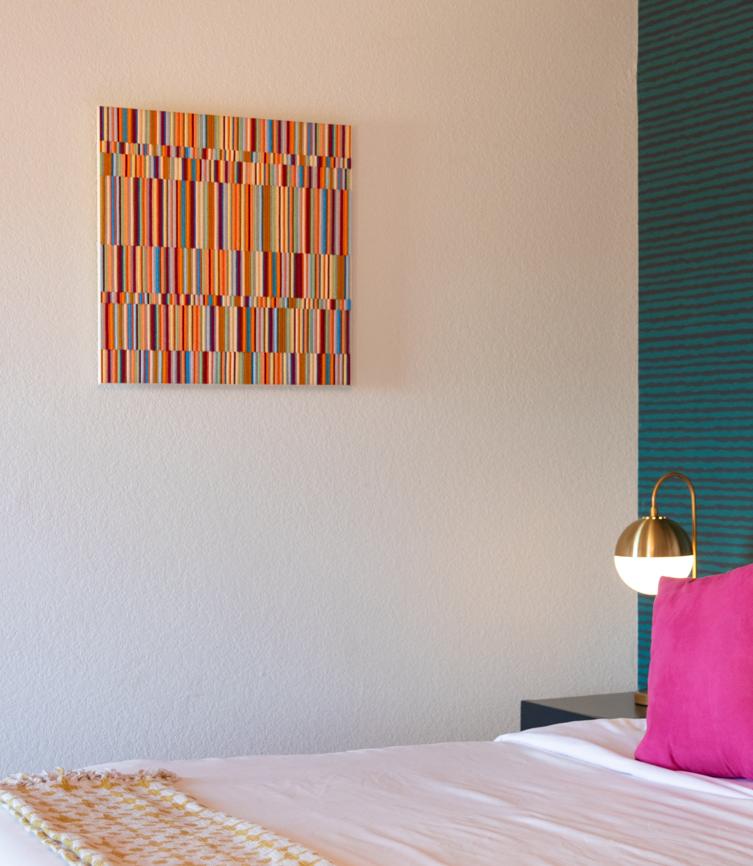 A square felt wall art panel hangs on the wall of a bedroom. It has a vibrant rainbow of felt colors on edge with alternating heights and various height rows. A patterned wallpaper, gold table lamp and pink pillow are seen at right.