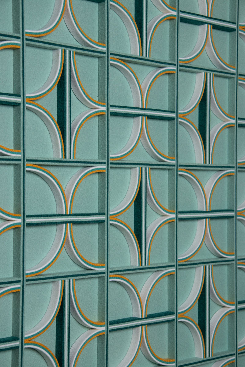 Patterned felt wall art with curved and straight felt details on edge. The background is a teal or sage color, and the details are yellow, white, teal, and dark green. 