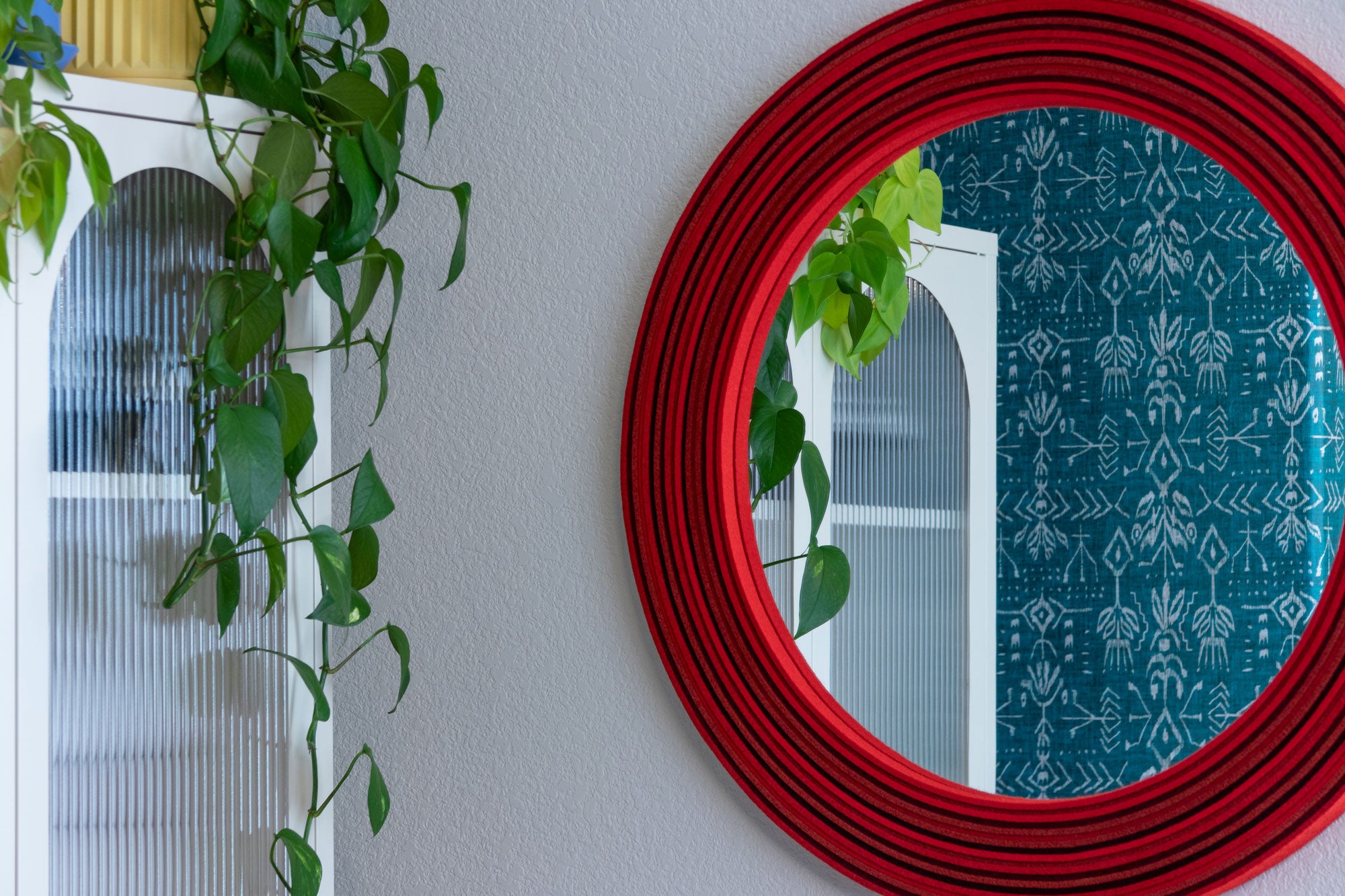 A large round mirror framed with multi-tone red felt hangs on a white wall. It reflects a white glass front storage cabinet, a drapping plant and  a blue patterned wallpaper. 