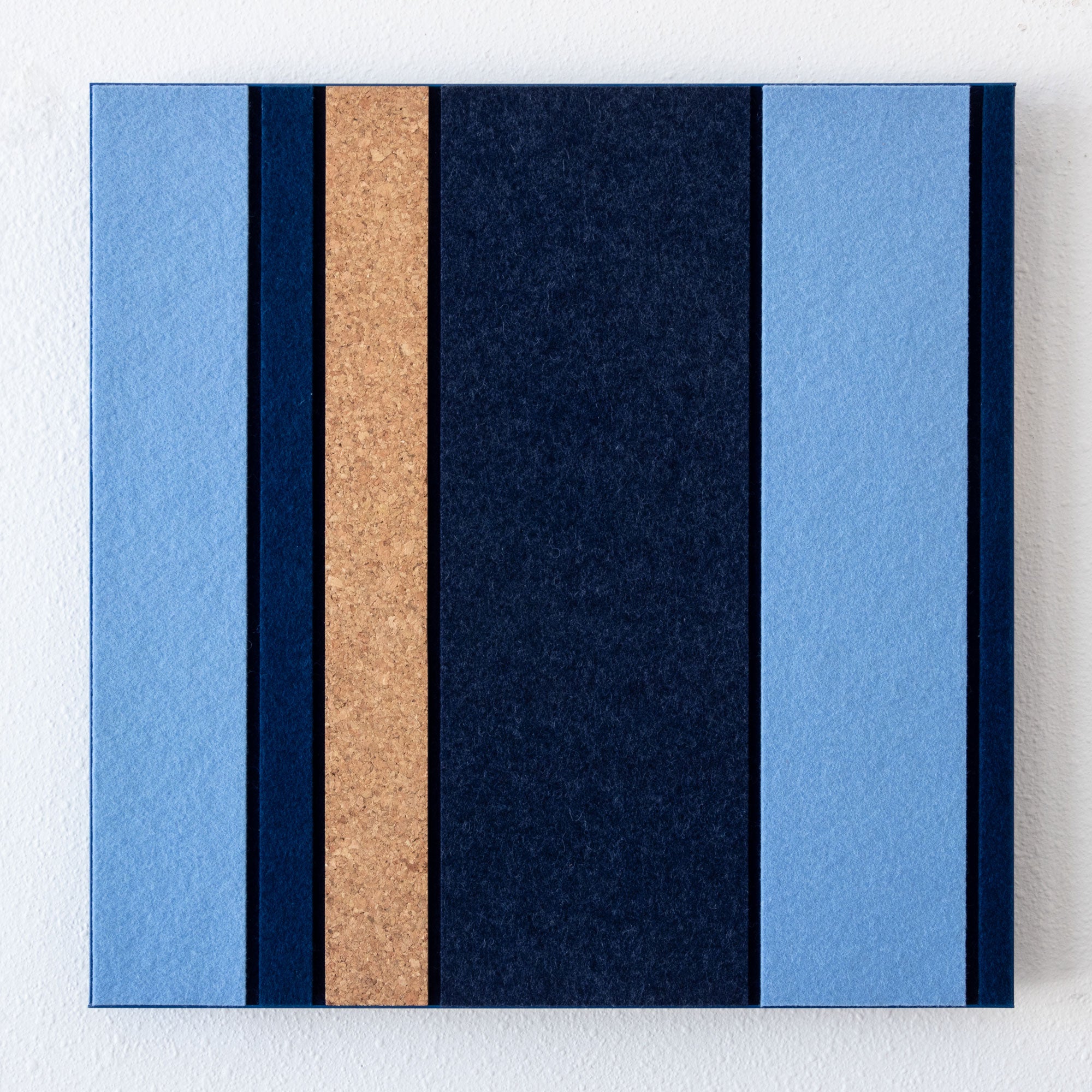 A square wall-mounted canvas or panel featuring vertical textured stripes in light blue, dark navy, and a tan cork-like center stripe, shown against a white wall.
#color_blues