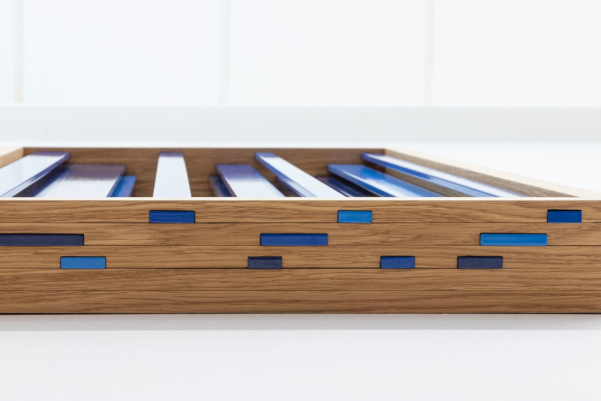 A close-up of the bottom of an abstract mirror, with blue metal louvers peeking through the wooden frame, set against a clean white background.