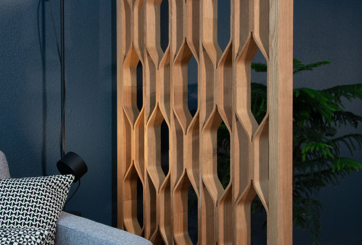 A tall wooden lattice-style screen with a geometric interlocking pattern, shown in a modern interior setting next to a sofa and a plant.