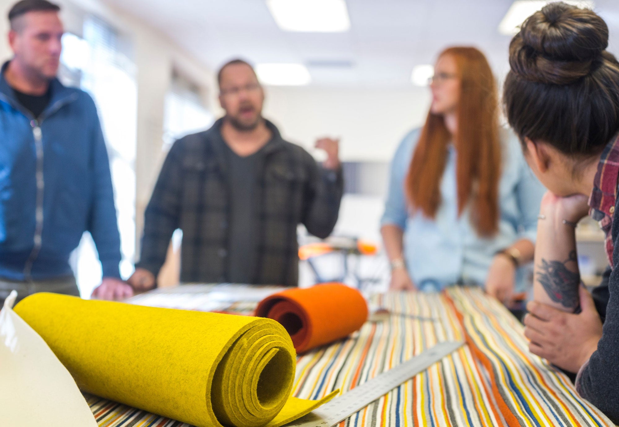 An out of focus shot people standing around a work table with colorful bolts and strips of felt. 