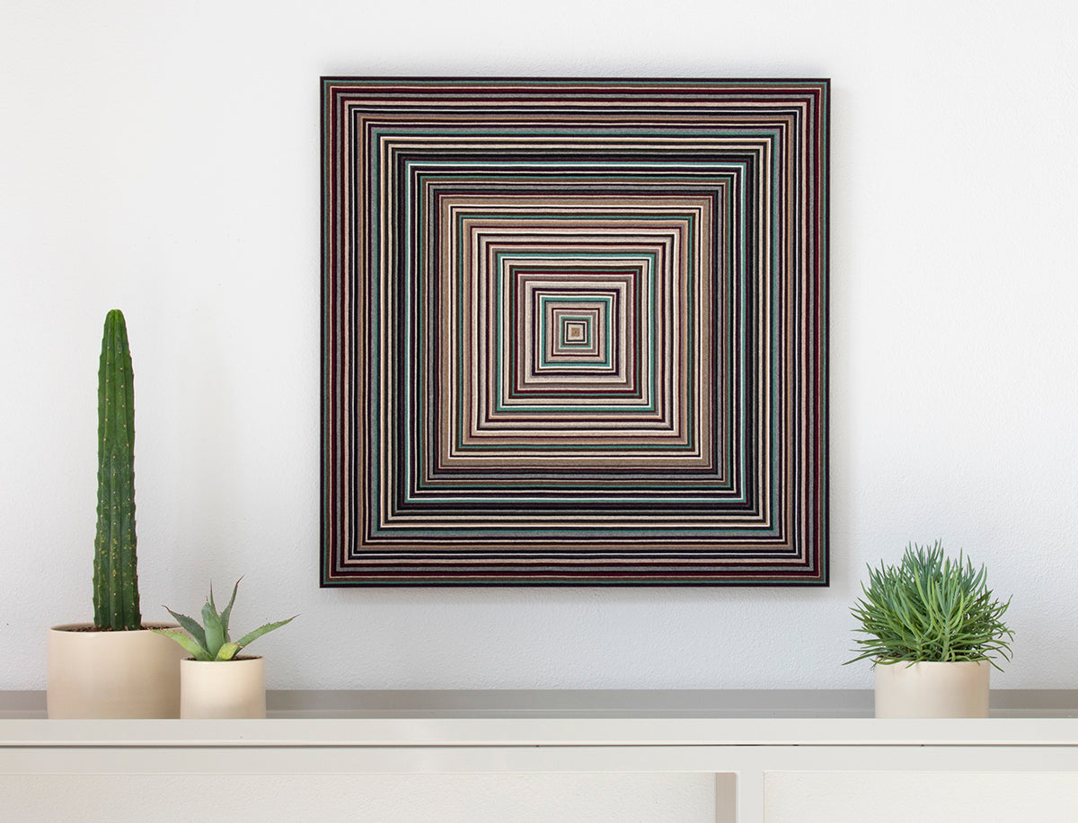 A felt wall panel of concentric squares of strips of wool felt in neutrals with a pop of red and turquoise. The square piece hangs on a white wall over a shelf with cactuses on it. #color_neutrals with teal