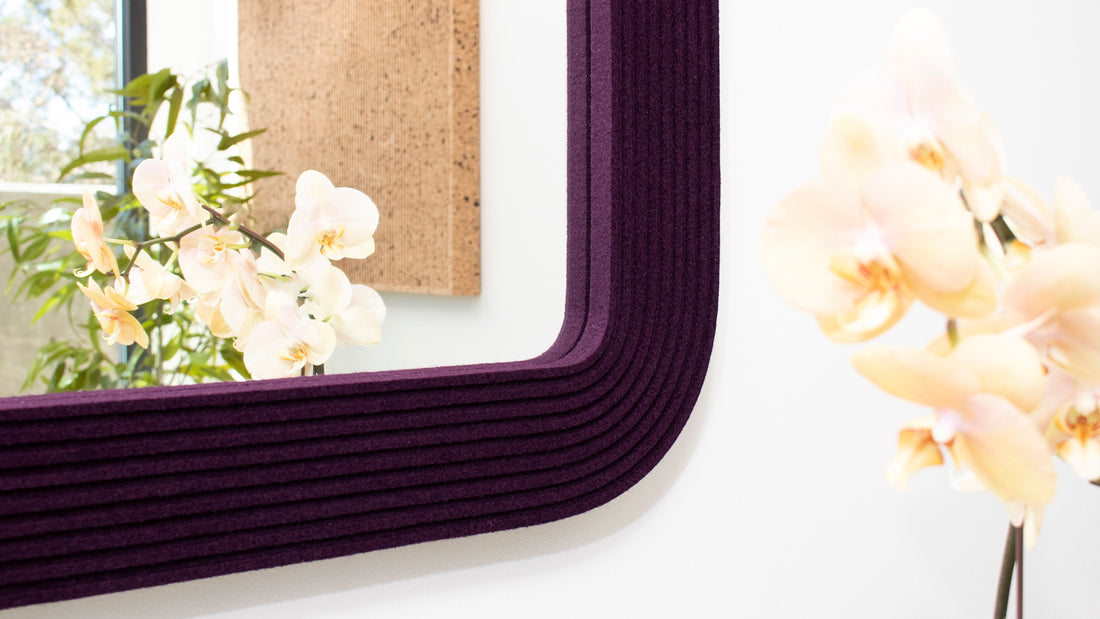 The corner of a rounded square mirror framed with eggplant colored felt stripes. An orchid is seen in the reflection