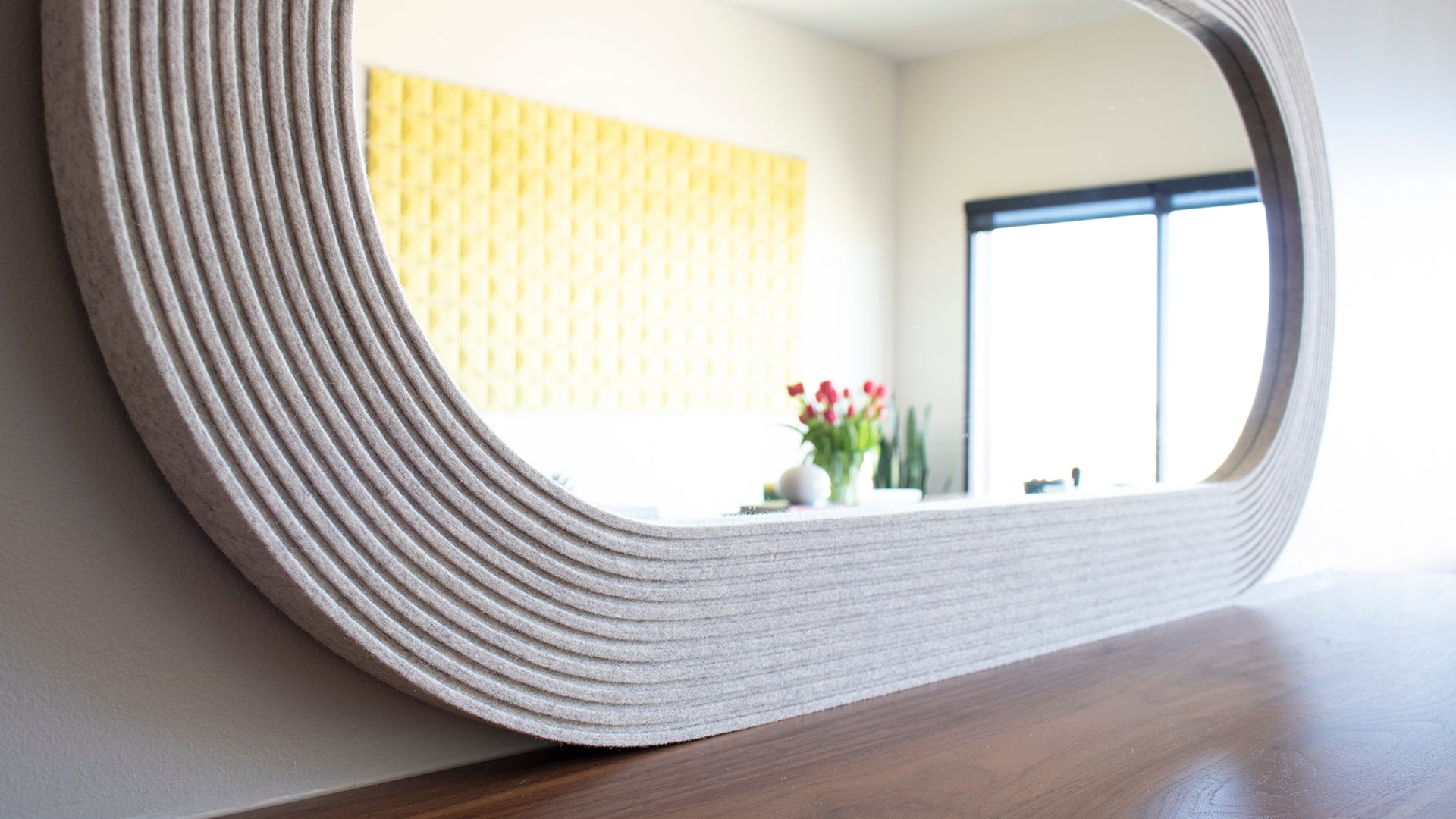 A light gray felt capsule shaped mirror sitting on a wood surface. The reflection shows a yellow wall art piece, pink tulips, and a large sliding door.