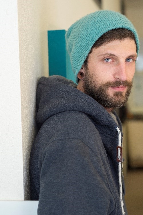 A man with a light blue beanie and a gray hoodie looks at the camera