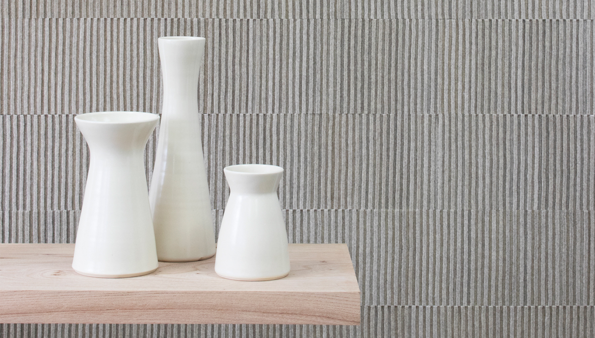 Ribbed wall covering in a gray heathered felt. In the foreground are three white ceramic vases on a wood shelf.