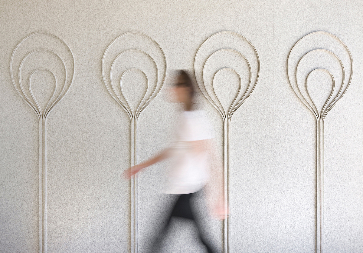 blurred person walks in front of Submaterial FilzFelt bulb wall covering in pale grey. The wall is designed with curvalinear whimsical bulb shapes.