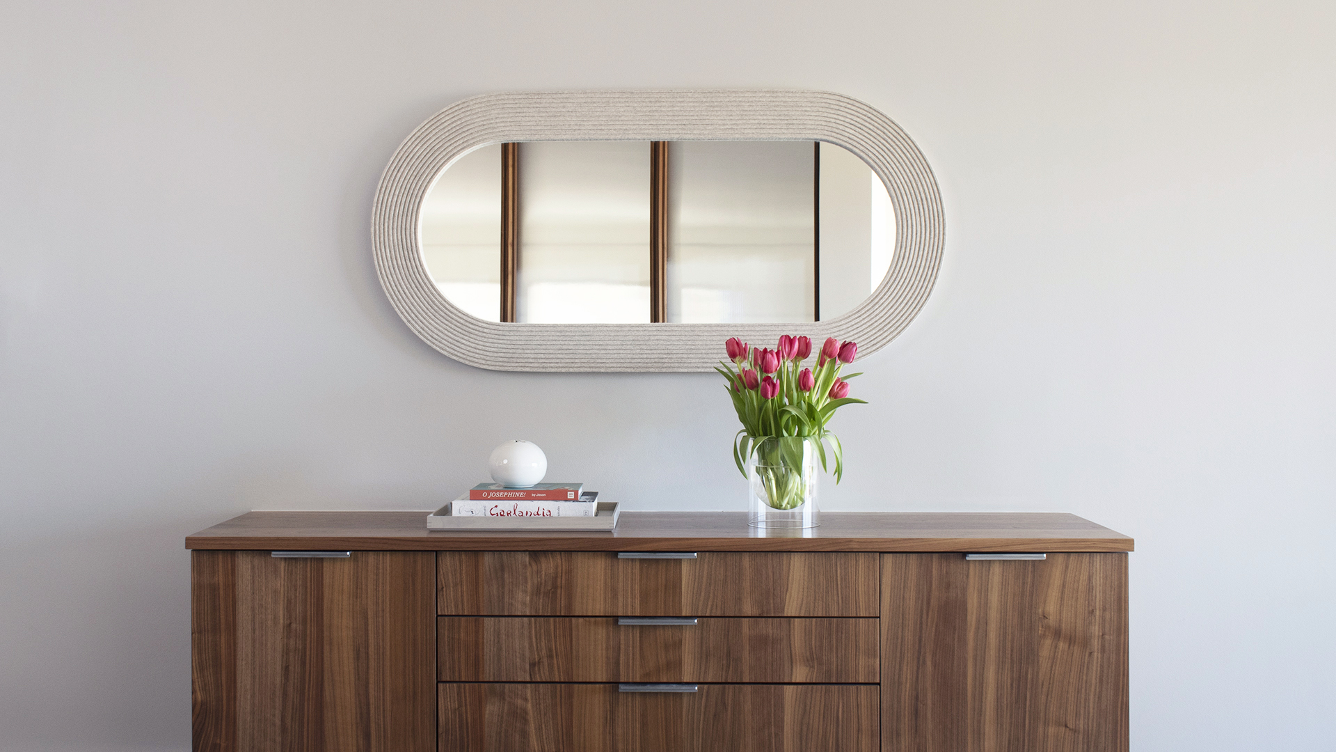 Pill shaped mirror in light gray felt hung horizontal over a sideboard with pink tulips