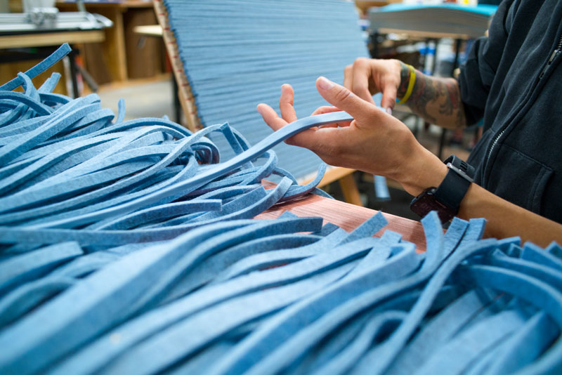The hands of a figure in a black zip up hoodie grab a long strip of blue felt from a pile spread out on a table.