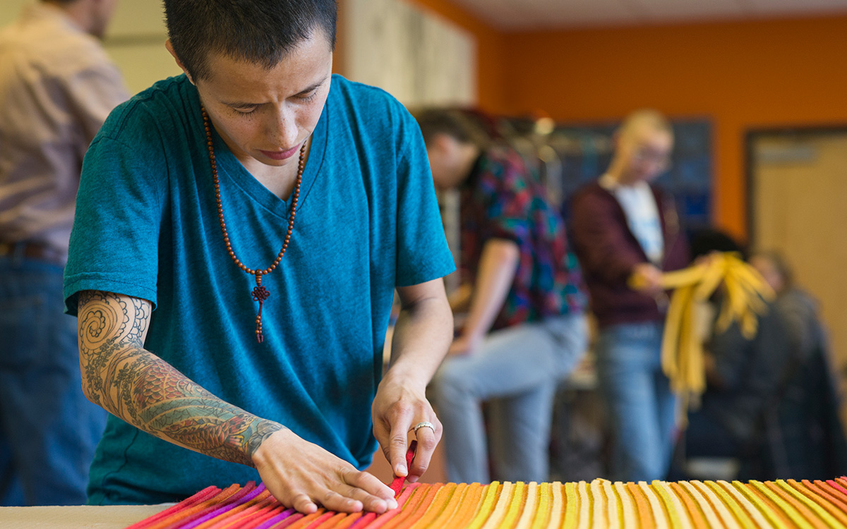 Fabricator in a blue shirt with short hair and tattoos works on a yellow, orange and pink felt panel