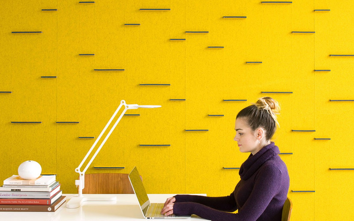Woman sits at a desk in front of a bright yellow felt wallwith blue horizontal tabs.
