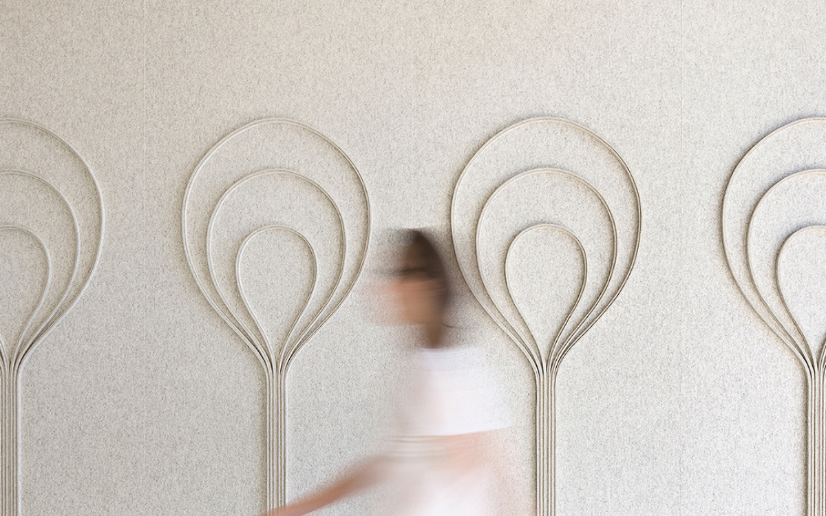 A figure is blurred by movement walking in front of a heathered white acoustic felt wall with blub shaped repeating details