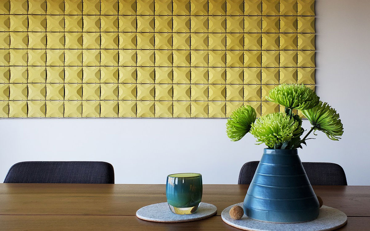 Yellow suede wall hanging with a 3-dimensional x-shaped pattern hung behind a dining table in a residential interior with a large blue vase and green flowers