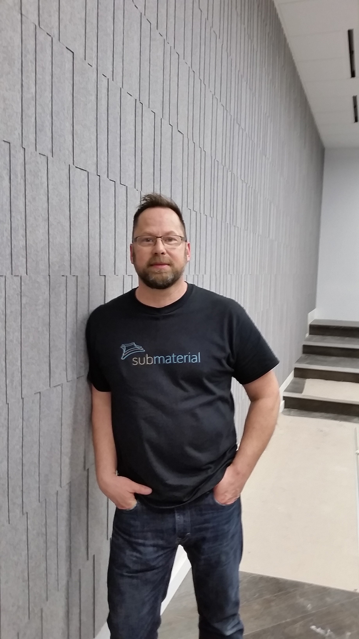 David Hamlin leaning against a gray felt wall covering wearing a t-shirt with the Submaterial logo.