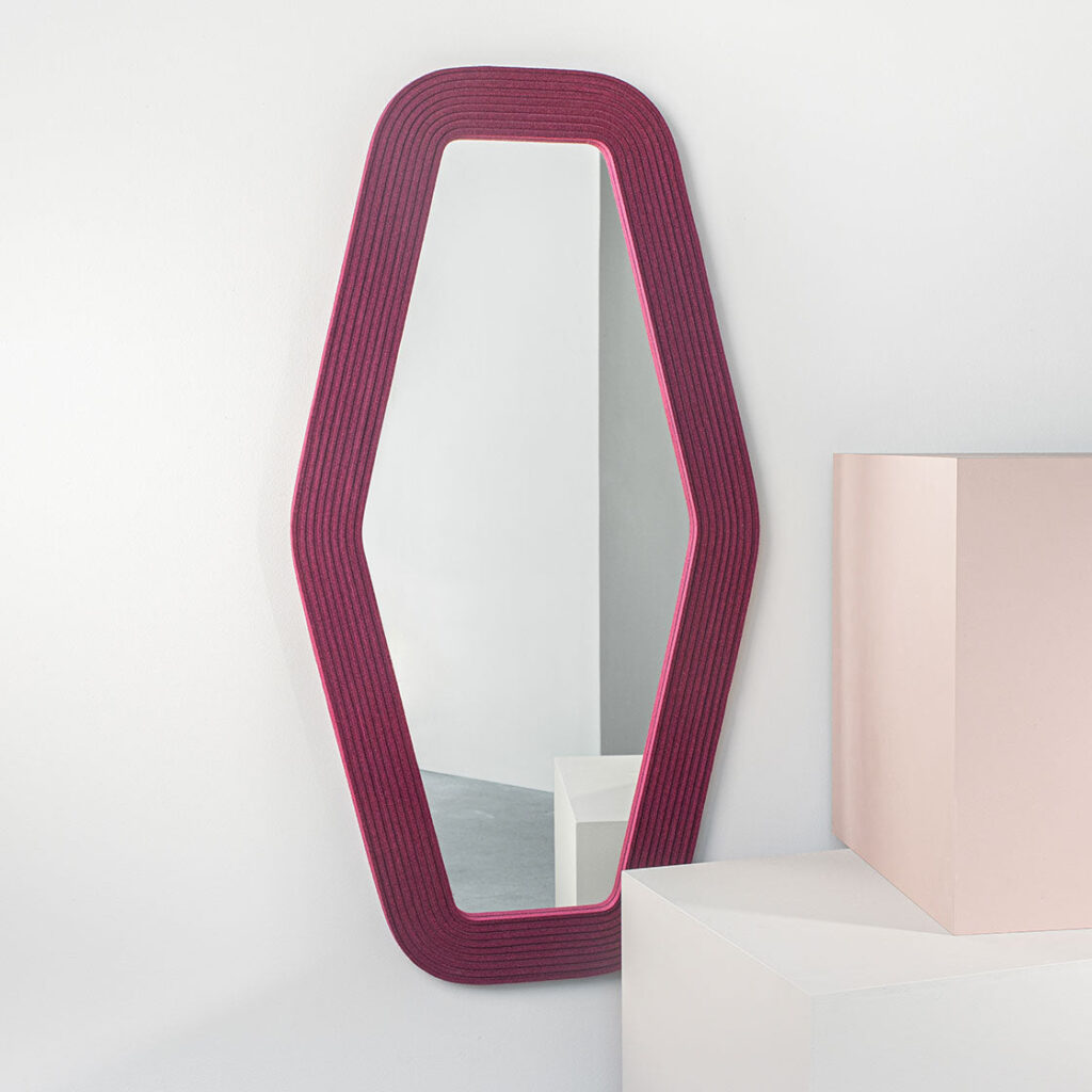 A hex-shaped mirror framed with deep red ribbed felt hung vertical next to a pink and white box