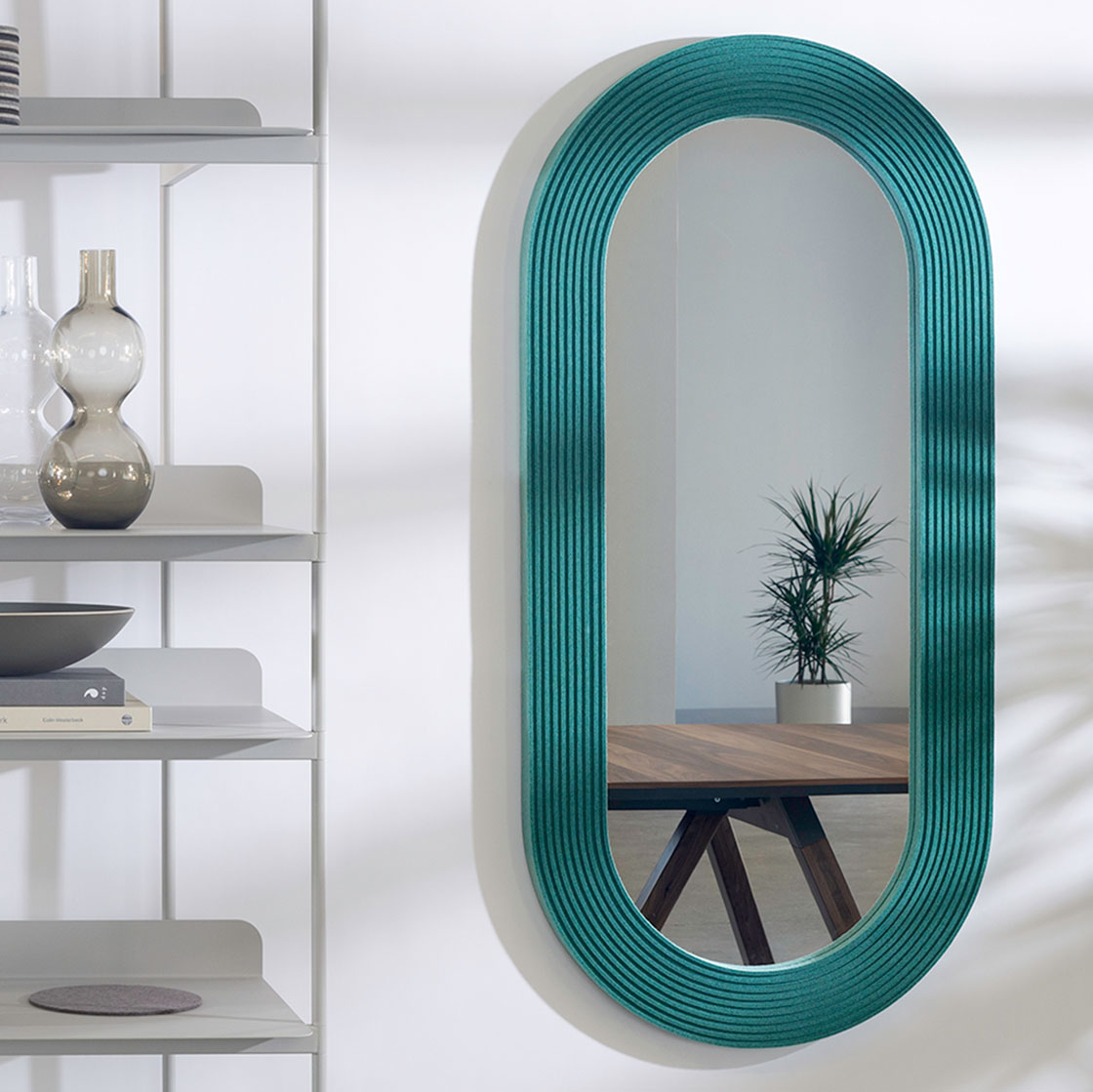 Pill shaped mirror framed with teal ribbed felt. It is reflecting a table and plant and is hung next to a shelving unit.