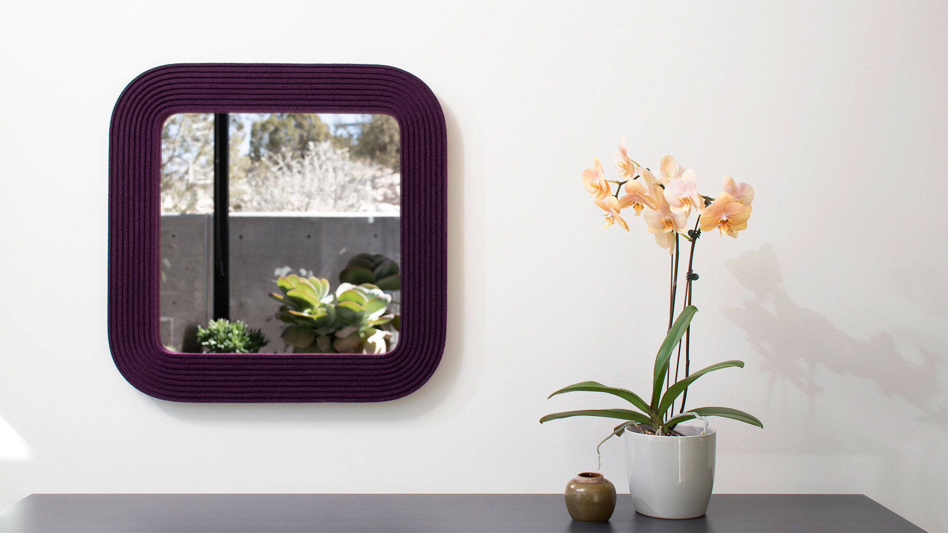 Wool felt square mirror in eggplant next to an orchid and plants reflected.